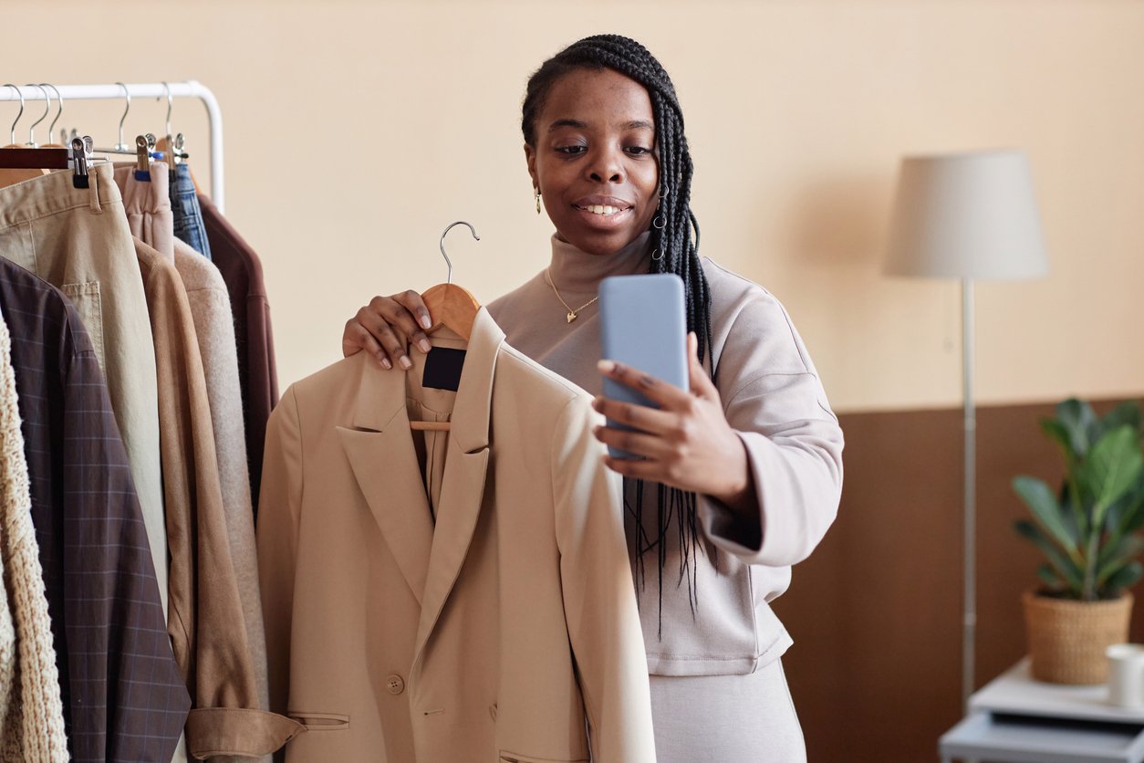 Woman holding suit jacket and looking at phone while stood besides clothes rail